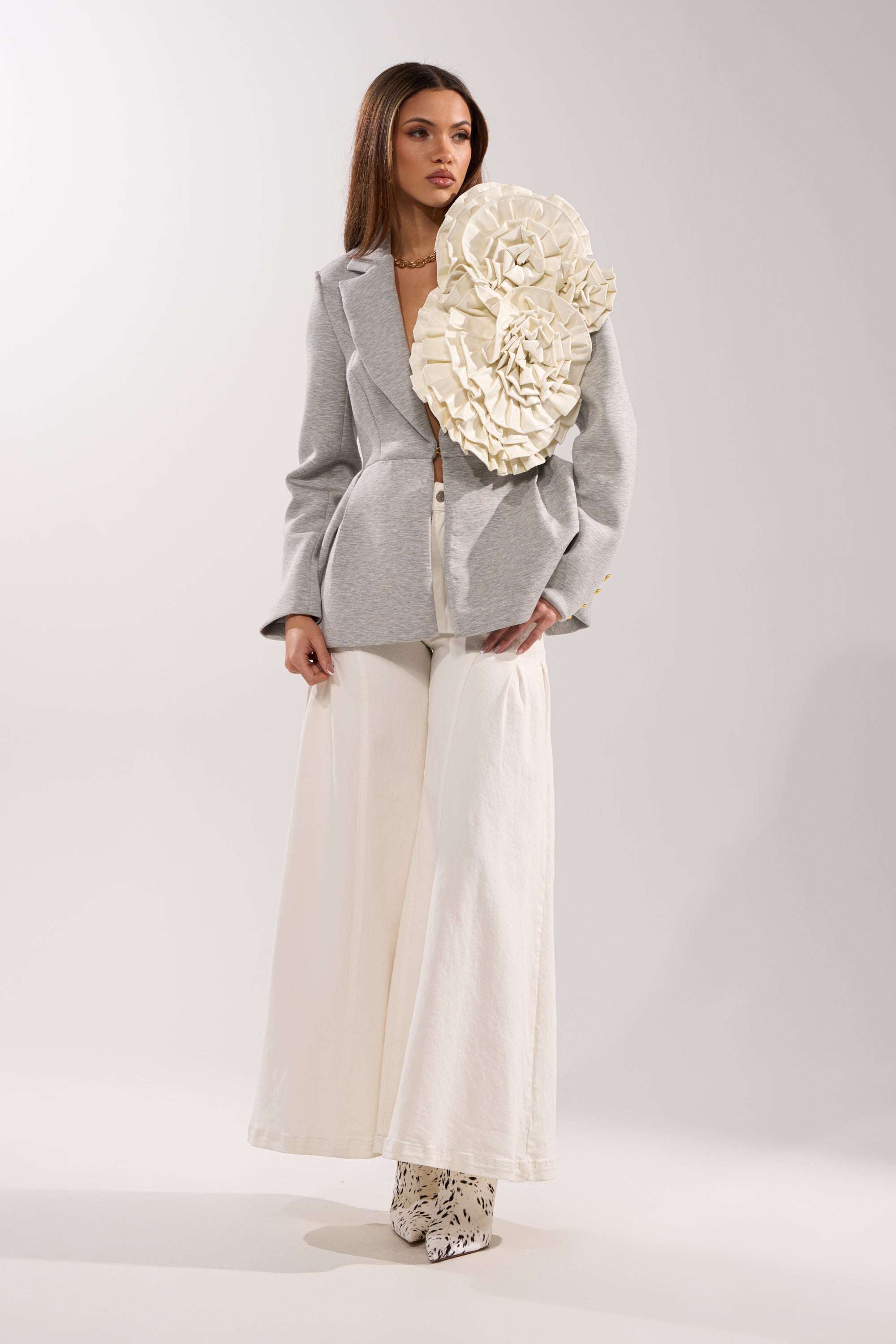A woman wears a light grey blazer featuring the BLOOMING PU OVERSIZED BROOCH as a statement, styled with wide-legged white pants and patterned white boots against a plain light background. 3
