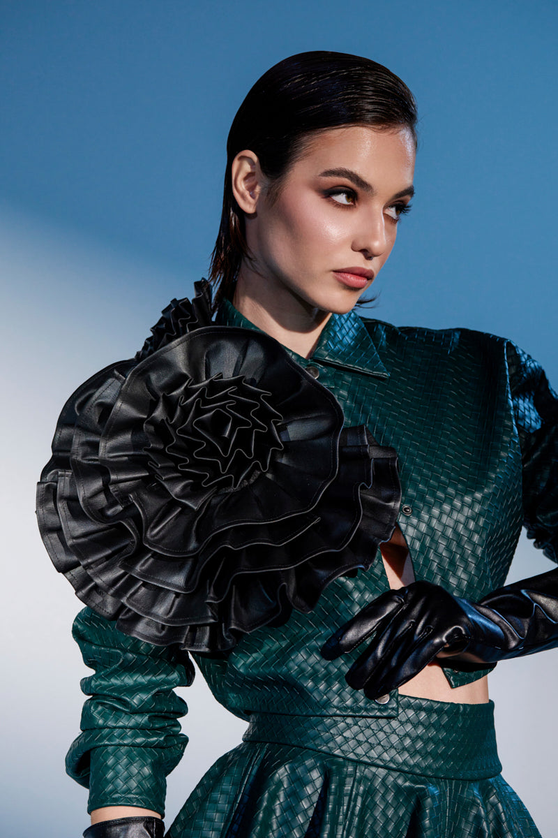 A woman poses against a blue gradient background wearing a textured green outfit, sleek side-parted hair, black leather gloves, and the FEELING ZESTY PU STATEMENT BROOCH—a large black ruffled floral accessory on her shoulder.