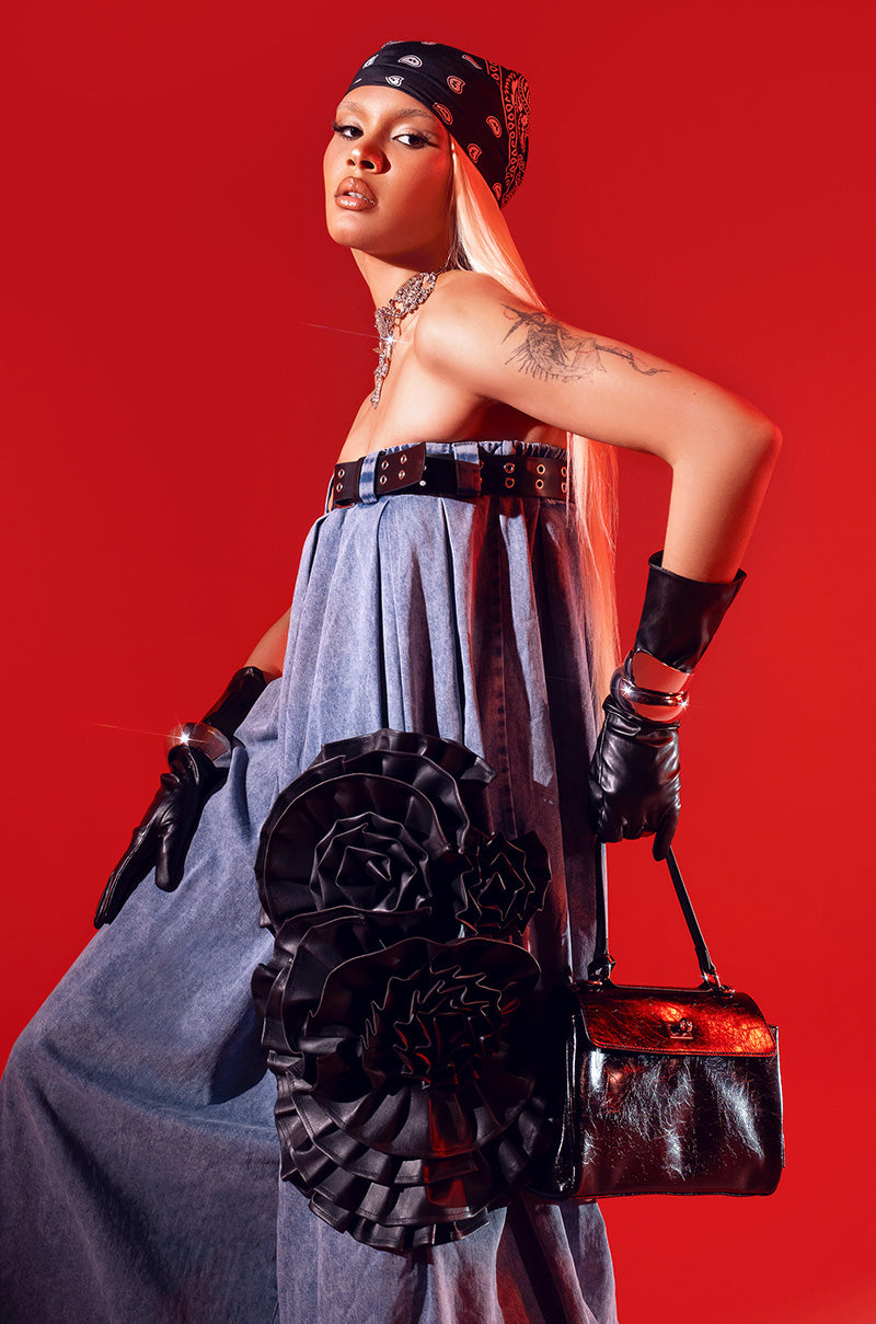 Person posing in front of a red background, wearing a strapless denim dress with ruffled floral details, black leather gloves, black bandana, silver choker, and holding the FEELING ZESTY PU STATEMENT BROOCH.