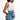 A woman in a sleeveless dress stands against a plain backdrop, wearing the MORE IS BETTER DENIM BROOCH with floral detailing at the waist. She has straight shoulder-length hair and poses with one hand on her hip.