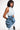 A woman in a sleeveless dress stands against a plain backdrop, wearing the MORE IS BETTER DENIM BROOCH with floral detailing at the waist. She has straight shoulder-length hair and poses with one hand on her hip.