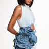 A woman in a sleeveless dress stands against a plain backdrop, wearing the MORE IS BETTER DENIM BROOCH with floral detailing at the waist. She has straight shoulder-length hair and poses with one hand on her hip.