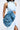 Dressed in a blue MORE IS BETTER DENIM BROOCH sleeveless cutout denim dress with a high collar, long skirt, and thigh-high slit, a person holds an oversized clutch with ruffled floral detailing. Both items seamlessly match in color.