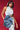 Against a red background, a woman wears a light blue sleeveless top and skirt, holding the MORE IS BETTER DENIM BROOCH. Her long earrings and the outfit's unique design add style with the large denim brooch featuring floral details.