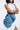 Wearing a sleeveless denim outfit with a high neckline and side slit, a person holds the MORE IS BETTER DENIM BROOCH, a large blue ruffled accessory with floral detailing. The oversized design stands out against the plain light background.