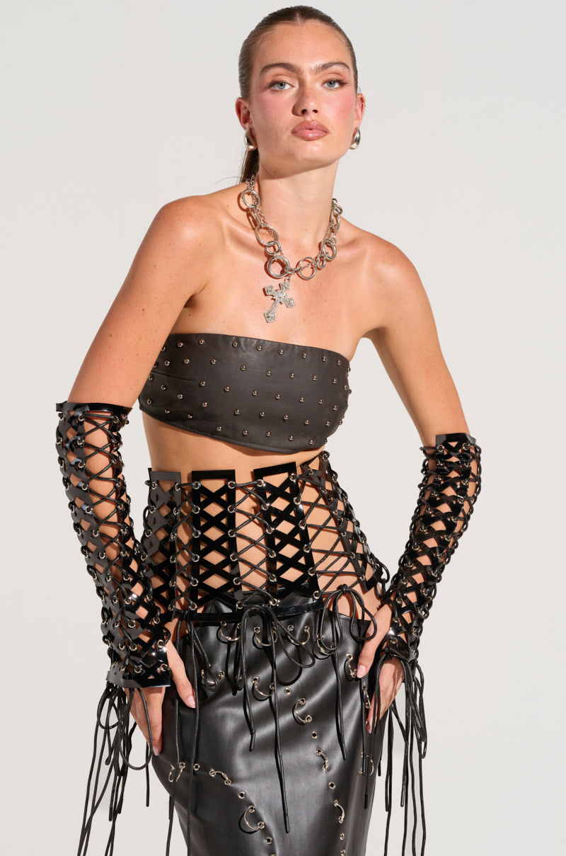 A person dressed in a black studded faux leather bandeau and skirt featuring a crisscross lace design, complemented by matching lace-up gloves. They sport the SO FEMME LACE UP BELT, accessorized with a silver chain necklace, standing with hands on hips against a neutral background.