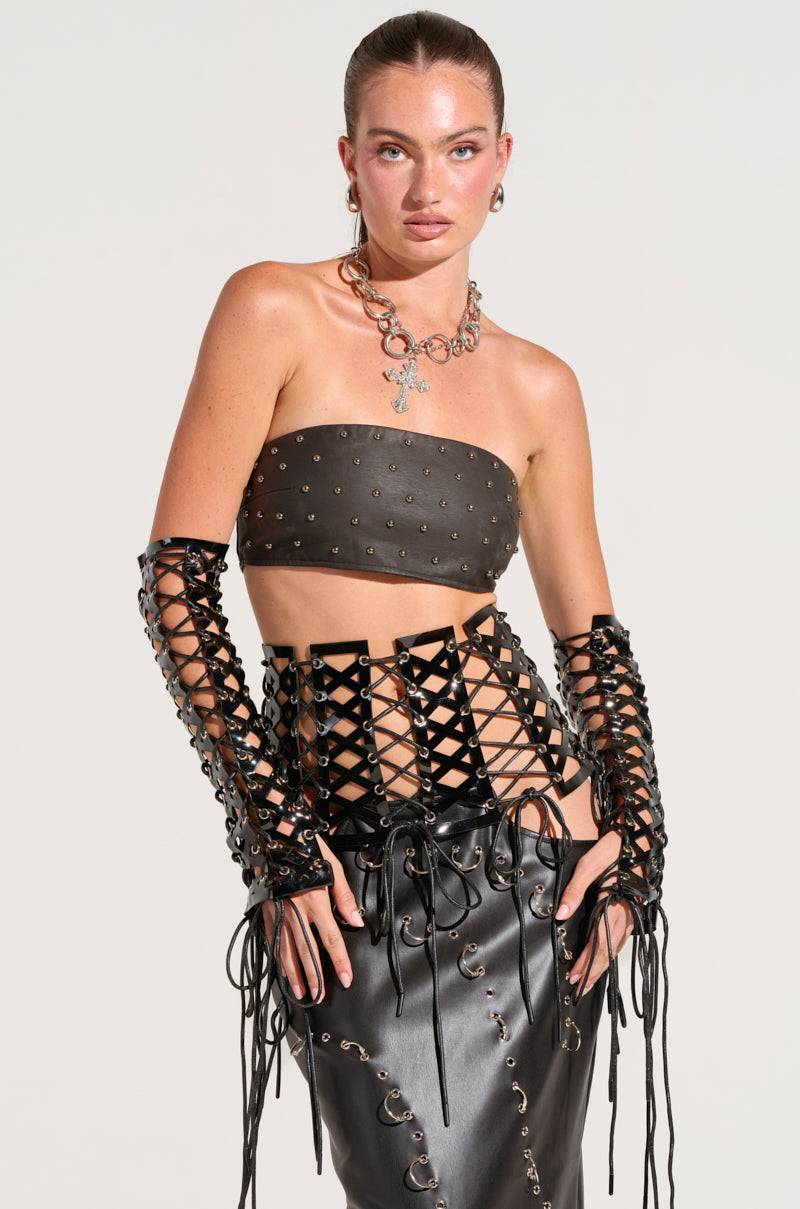 A person stands confidently in a black, strapless top and matching high-waisted skirt adorned with metallic rings. Their long, faux leather lace-up gloves hug their arms, complemented by a chunky silver necklace. A neat bun crowns their head, with the ensemble cinched together by the SO FEMME LACE UP BELT.