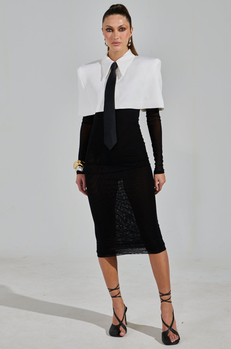 A woman confidently poses in the "She Means Business Tie Detail Mesh Midi Dress," featuring a large white collar and black tie. She accessorizes with strappy heels and gold bracelets, her hair slicked back, creating a poised look against a plain background.