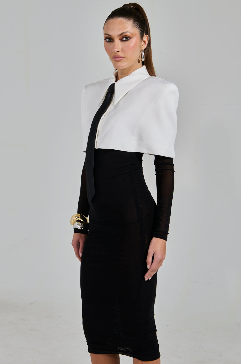 A woman with long brown hair in a high ponytail stands against a neutral background, wearing the SHE MEANS BUSINESS TIE DETAIL MESH MIDI DRESS featuring fitted black sleeves and complemented by a white cropped blazer, black tie, and large gold bracelet.