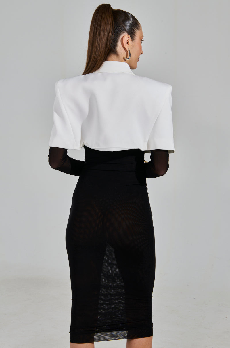 A woman with a high ponytail faces away, wearing the SHE MEANS BUSINESS TIE DETAIL MESH MIDI DRESS. It's a black, sheer bodycon dress with adjustable tank straps that falls below her knees. She pairs it with a cropped blazer against a plain light gray background.