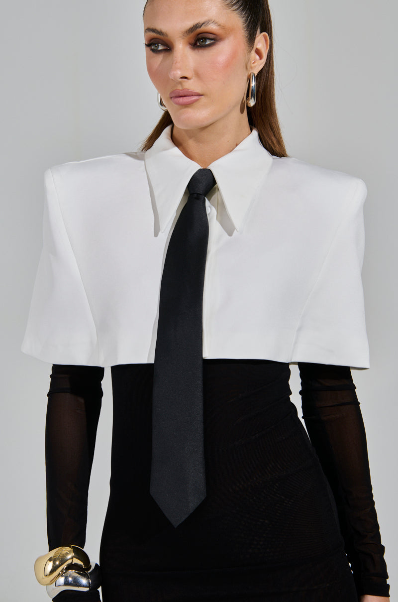 Wearing the SHE MEANS BUSINESS TIE DETAIL MESH MIDI DRESS, a person with long brown hair tied back sports a white cropped blazer and black tie. Large hoop earrings and a gold bracelet adorn their wrist against a plain gray background.