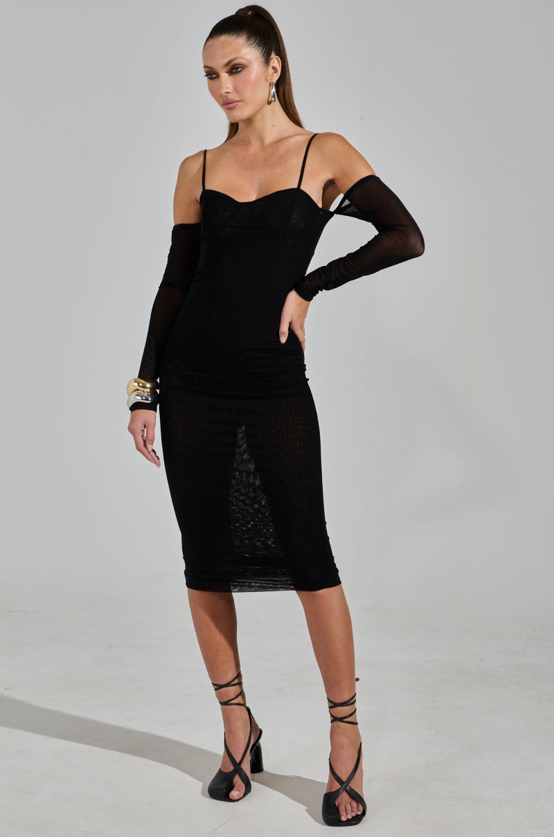 A person poses confidently in the SHE MEANS BUSINESS TIE DETAIL MESH MIDI DRESS with spaghetti straps and sheer long sleeves, complemented by strappy black heels and gold hoop earrings. Their sleek ponytail finishes the look.