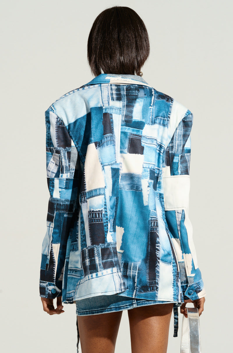 Person standing with their back to the camera, wearing the BLUE JEAN BABY DENIM PRINT BLAZER. The oversized jacket is crafted from a patchwork of different denim fabrics in various shades of blue, featuring long sleeves and falling to the upper thigh. The setting includes a plain, light-colored wall in the background.