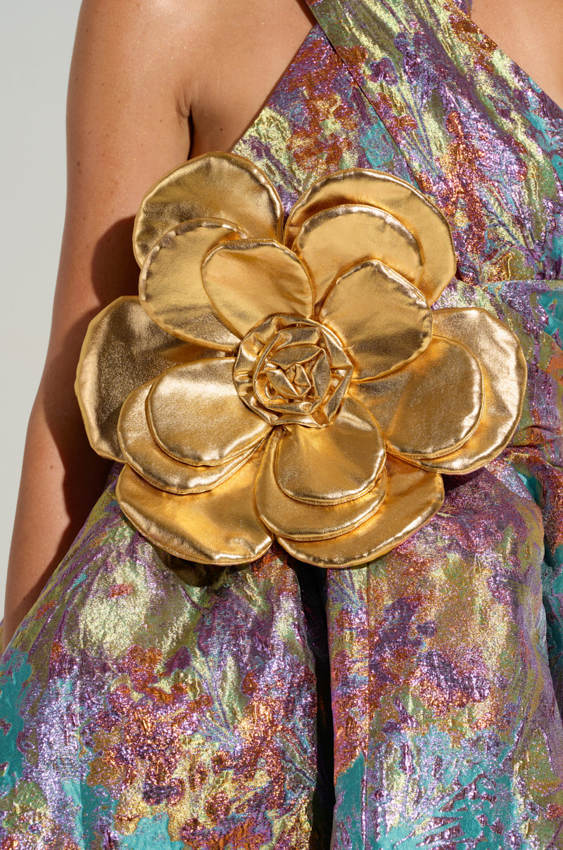 Close-up of a person wearing a colorful, shimmering fabric dress in a blend of green, purple, and pink hues. The dress features the GOLDEN HOUR METALLIC BROOCH as an accent. It has a textured, iridescent finish and intricate detailing with a discreet safety pin fastening.