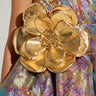 Close-up of a person wearing a colorful, shimmering fabric dress in a blend of green, purple, and pink hues. The dress features the GOLDEN HOUR METALLIC BROOCH as an accent. It has a textured, iridescent finish and intricate detailing with a discreet safety pin fastening.