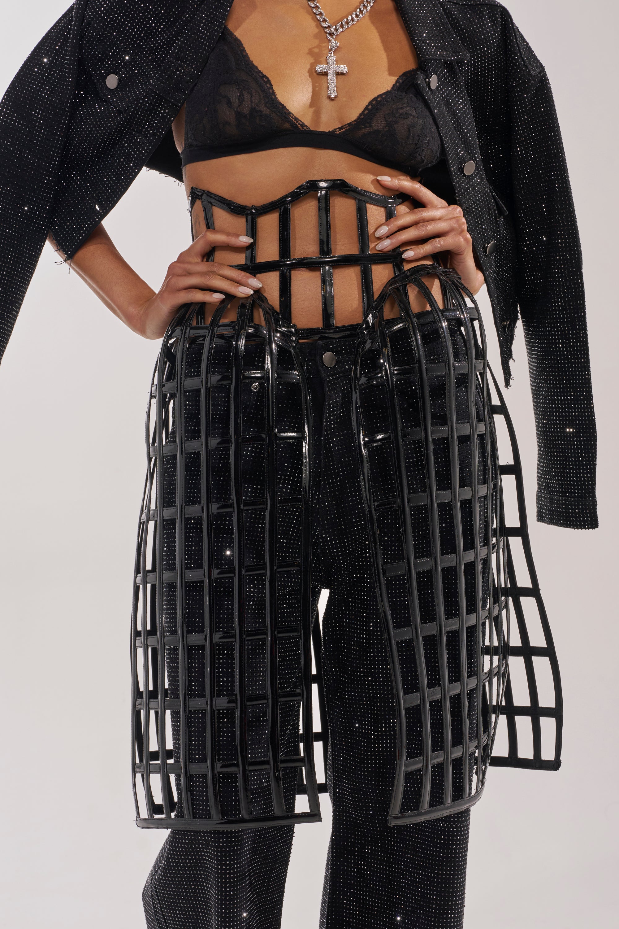 A person in a sparkly black bra, crop jacket, and pants poses with hands on hips, wearing the WILD THANG CAGE BELT over their waist and hips, accessorized with a large cross necklace. 0