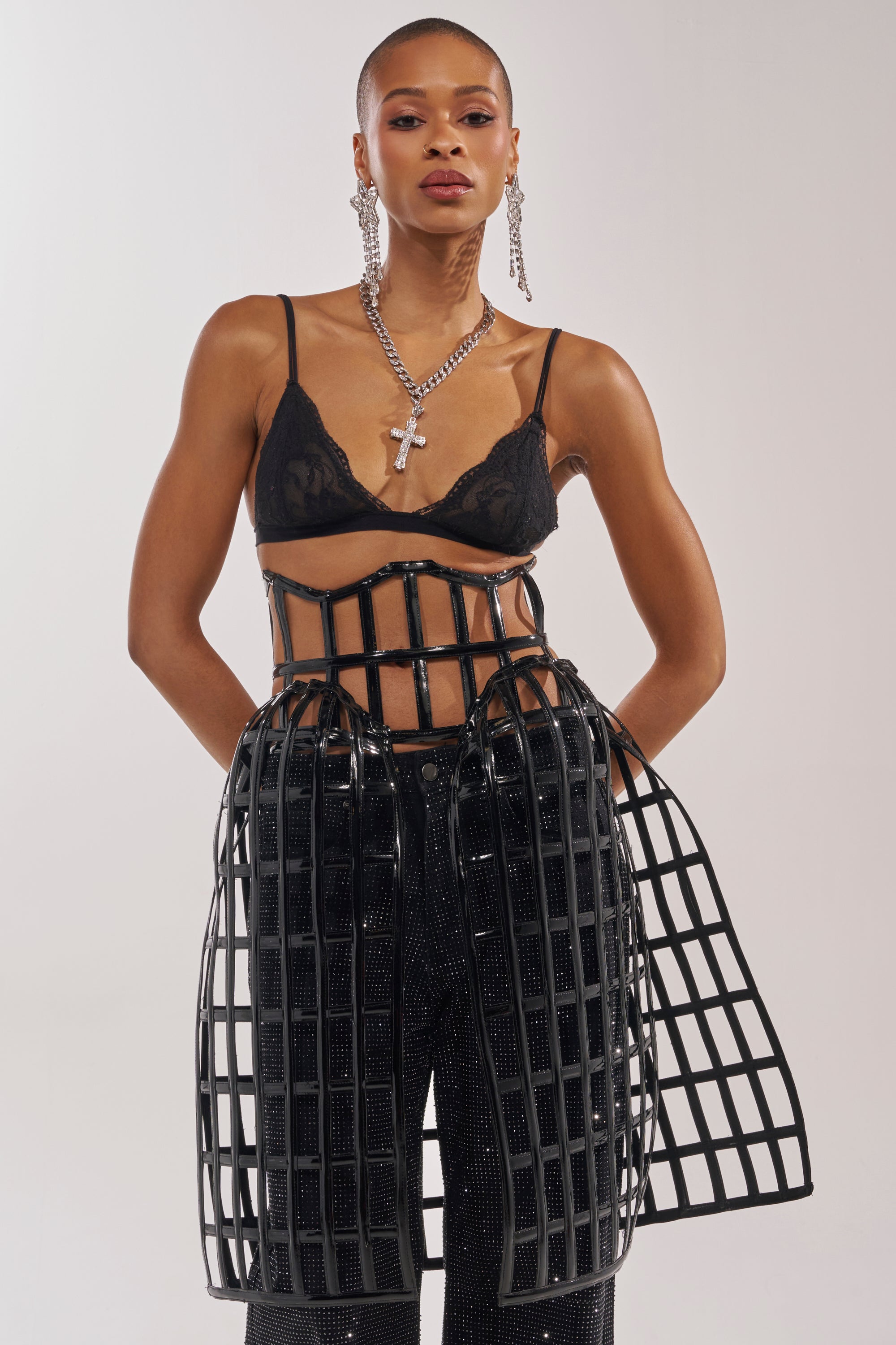 A model stands confidently against a plain light background wearing sparkly black pants and the WILD THANG CAGE BELT styled over a black lace bralette, accessorized with layered necklaces and long earrings. 1