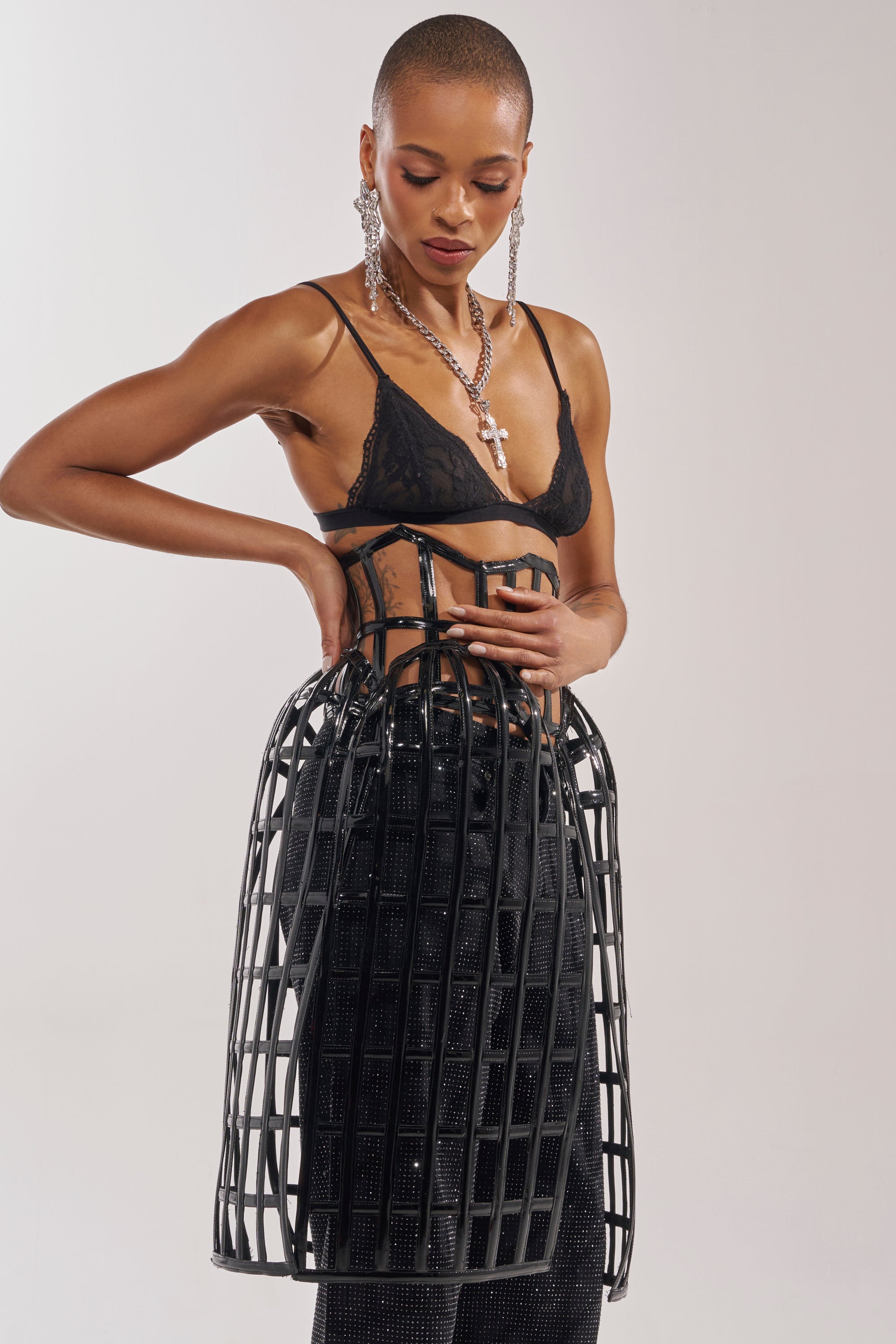 A woman styles the WILD THANG CAGE BELT—a black faux leather statement accessory—over a black lace bralette and sparkly pants, adding bold earrings for an avant-garde look against a plain background. 2