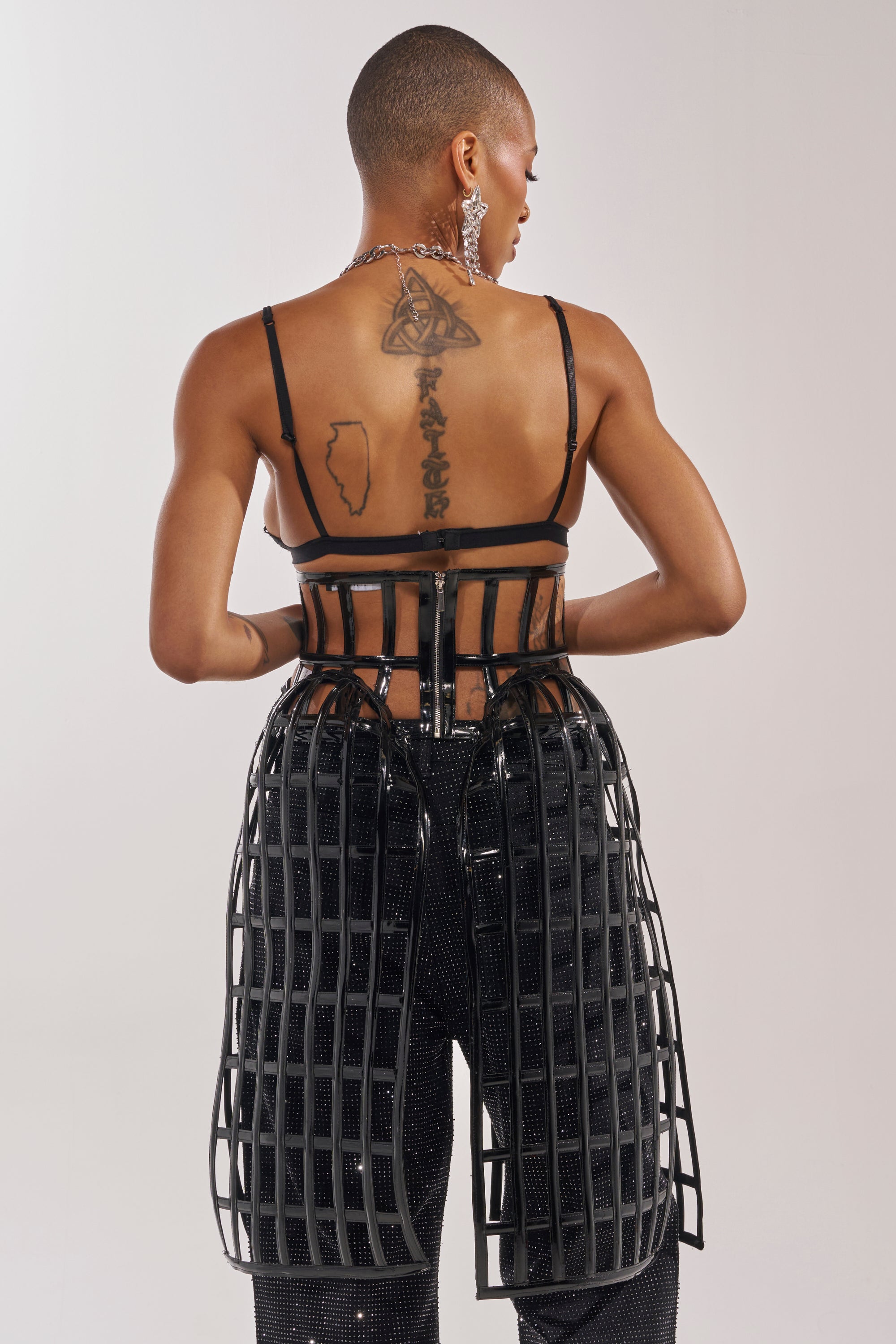 Photographed from behind against a plain background, a person with a shaved head and back tattoos wears the WILD THANG CAGE BELT over sequined pants, capturing an avant-garde fashion vibe. 3