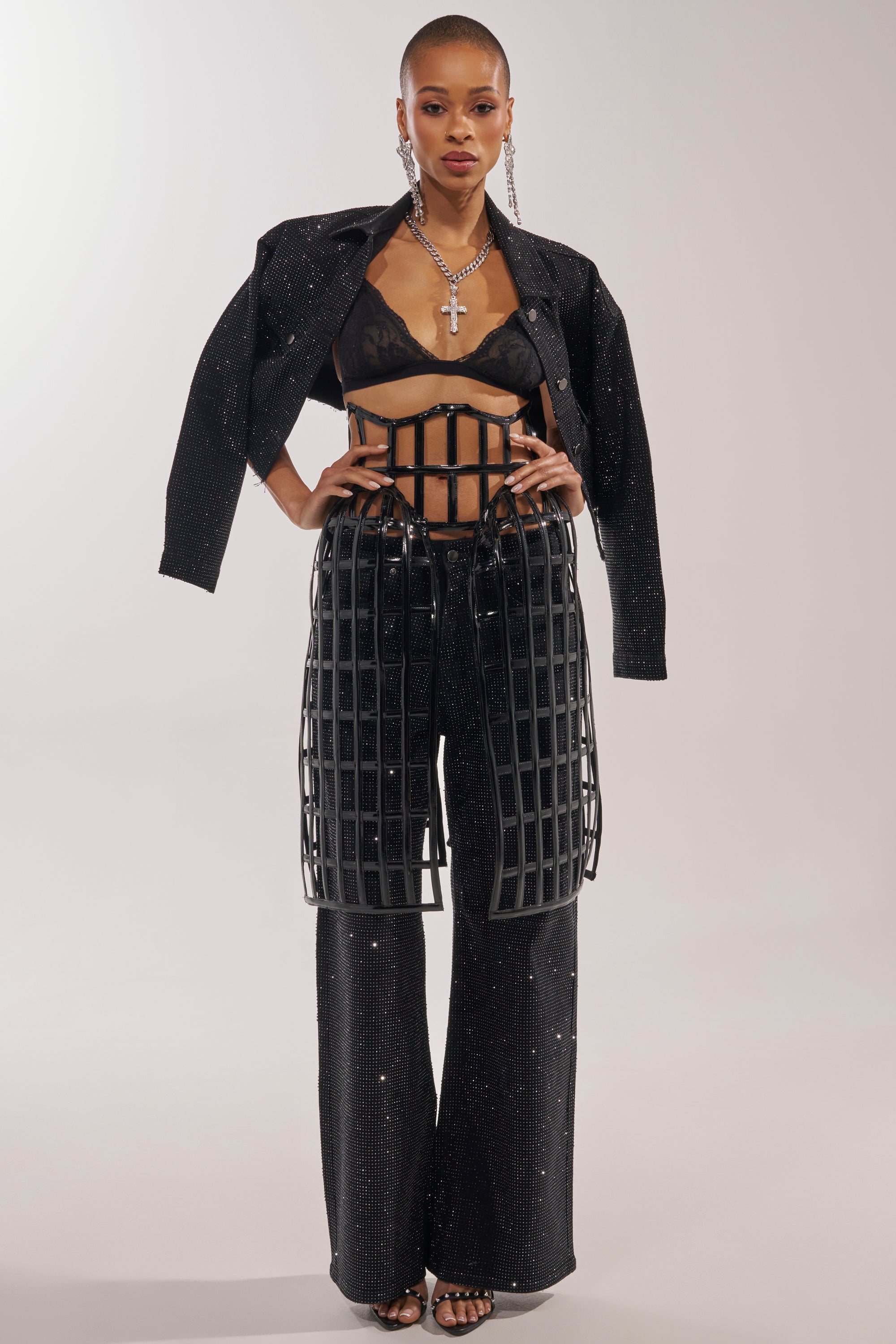 A woman poses confidently in a sparkling black outfit featuring the WILD THANG CAGE BELT, wide-leg pants, sheer black bra, cropped jacket, statement earrings, a bold cross necklace, and strappy high heels. 4