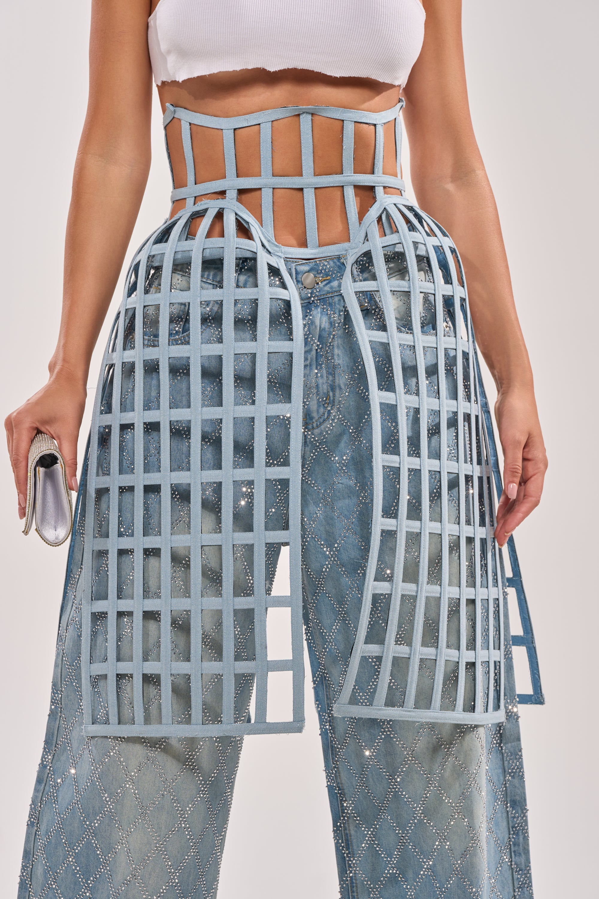Wearing a white crop top, embellished blue jeans, and the NEVER CAGED BELT—a denim grid-patterned skirt overlay extending from waist to mid-thigh—while holding a small silver clutch. 0