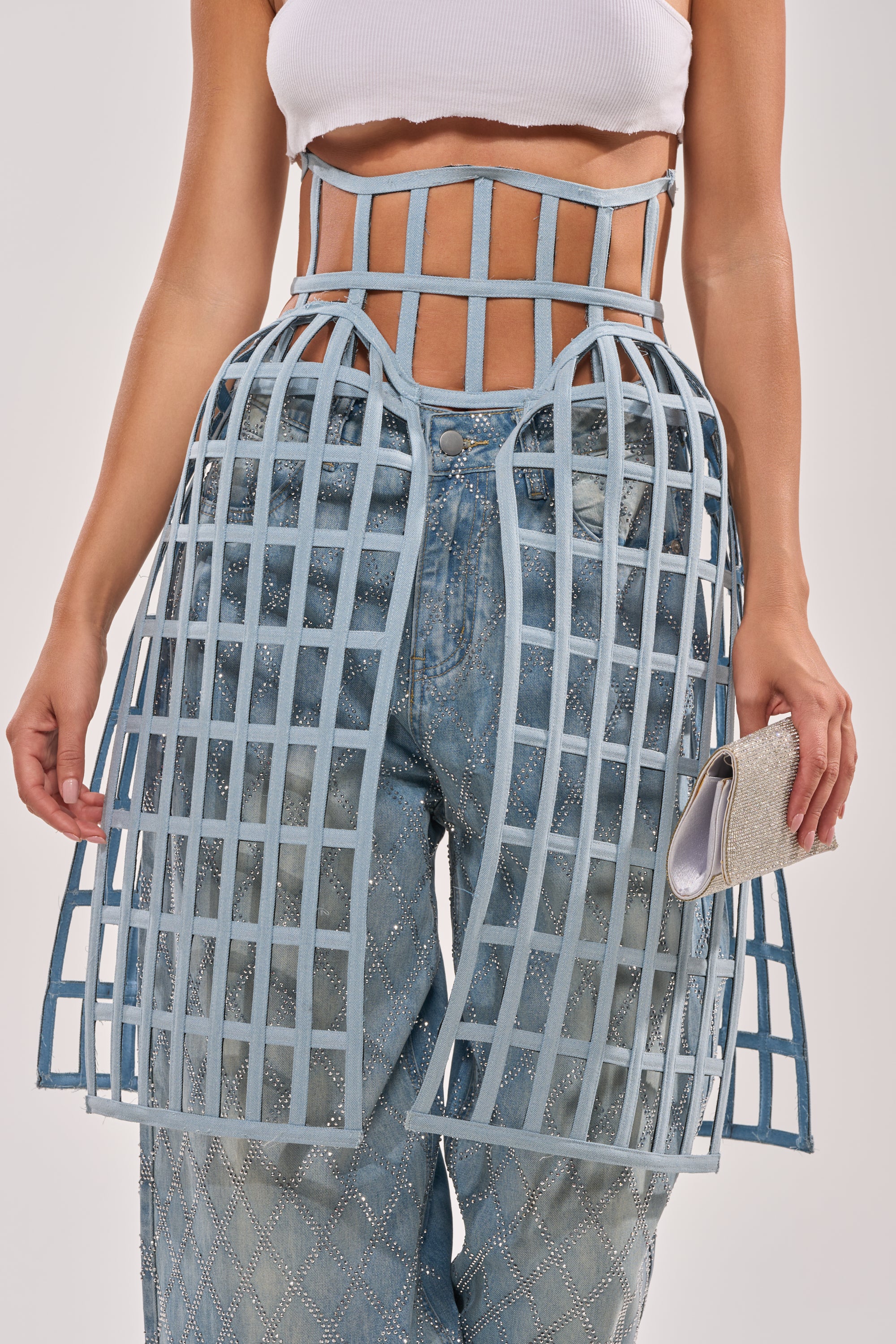 A woman dons a white crop top, blue diamond-pattern jeans, and the NEVER CAGED BELT—a light blue denim cage overlay—paired with a silver clutch for an avant-garde look. 2