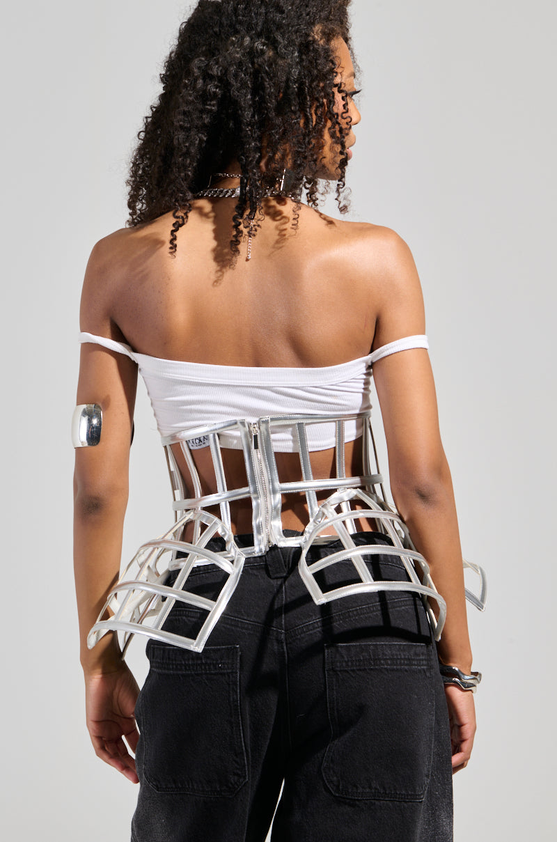 A person with curly hair is wearing a white off-shoulder top paired with the ULTIMATE TEASE METALLIC CAGE BELT around their waist. The cropped fit enhances their black pants, complemented by a silver arm cuff, as they face away from the camera.