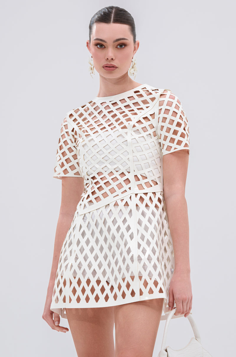 A woman stands against a plain background wearing the SHE'S DIFFERENT Cut Out Faux Leather Mini Dress in White. The short mini dress features a grid pattern, sheer sections, and short sleeves. She accessorizes with statement earrings and a white handbag.