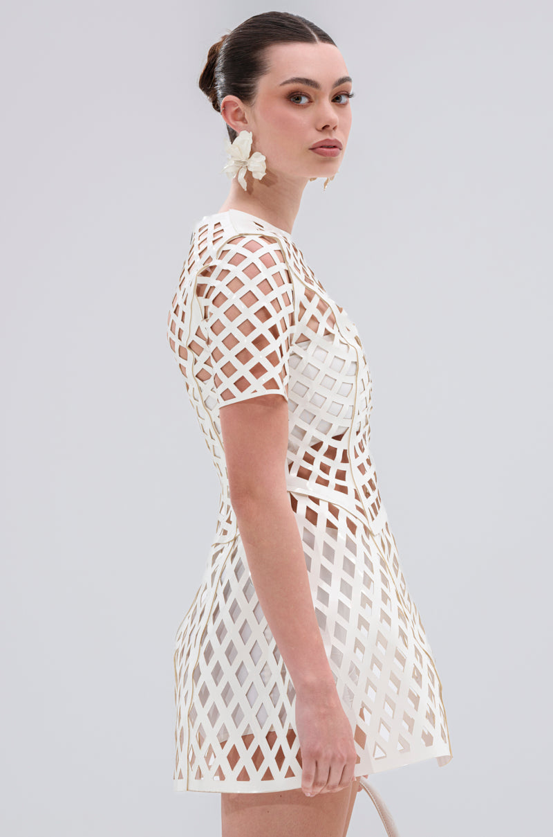A woman stands sideways, looking over her shoulder, wearing the SHE'S DIFFERENT CUT OUT FAUX LEATHER MINI DRESS IN WHITE with a bun hairstyle and large white flower earrings.