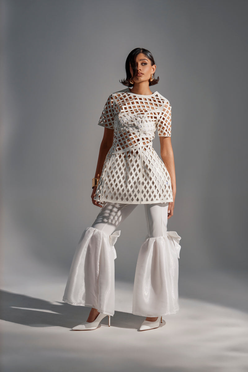 A woman models the SHE'S DIFFERENT CUT OUT FAUX LEATHER MINI DRESS IN WHITE, paired with dramatic white pants and geometric accents, accessorized with white heels and gold jewelry against a gray backdrop.
