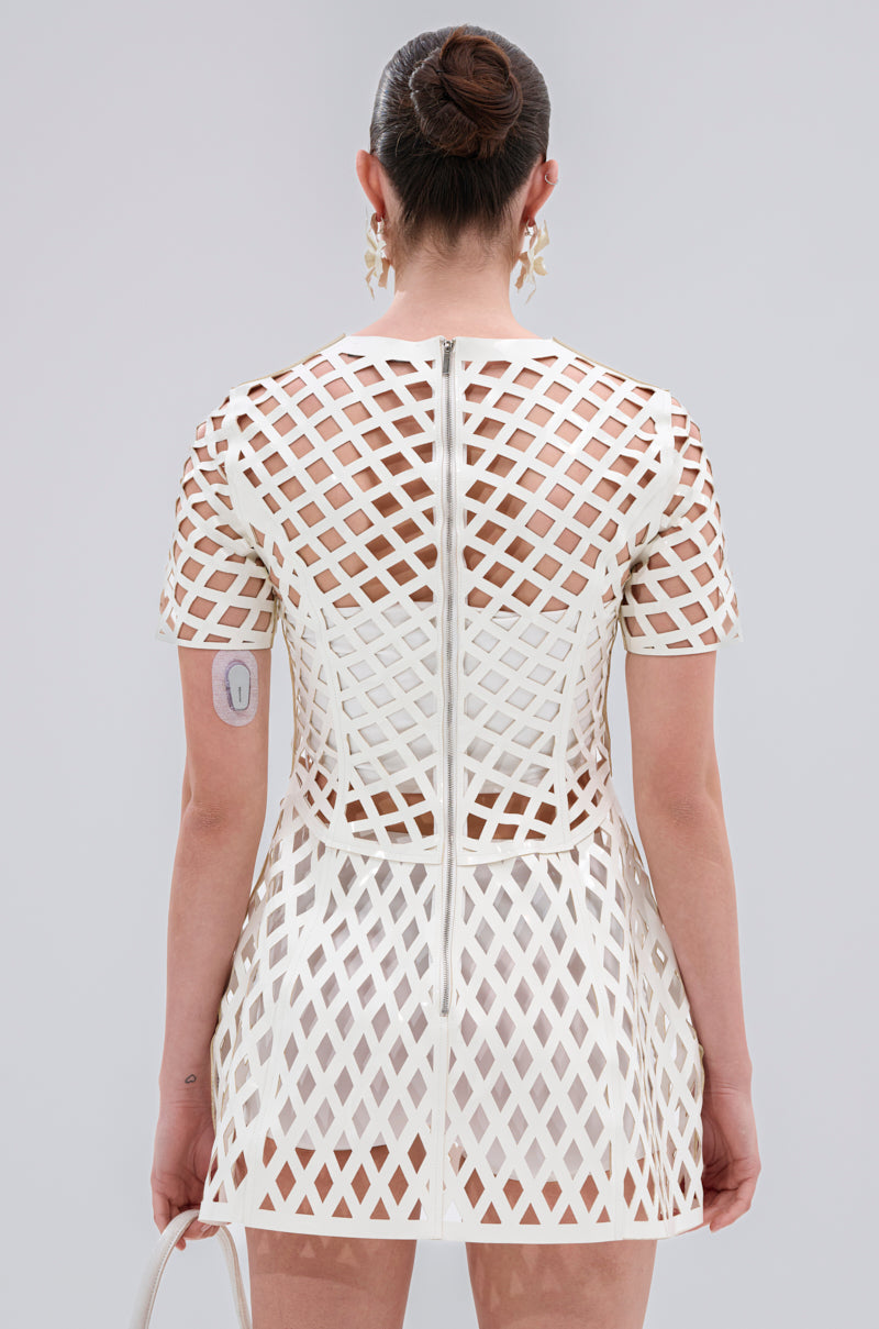 A person with dark hair in a neat bun is seen from behind wearing the SHE'S DIFFERENT CUT OUT FAUX LEATHER MINI DRESS IN WHITE, featuring geometric lattice cut-outs that reveal skin. They hold a white bag and have an arm patch on their left arm.