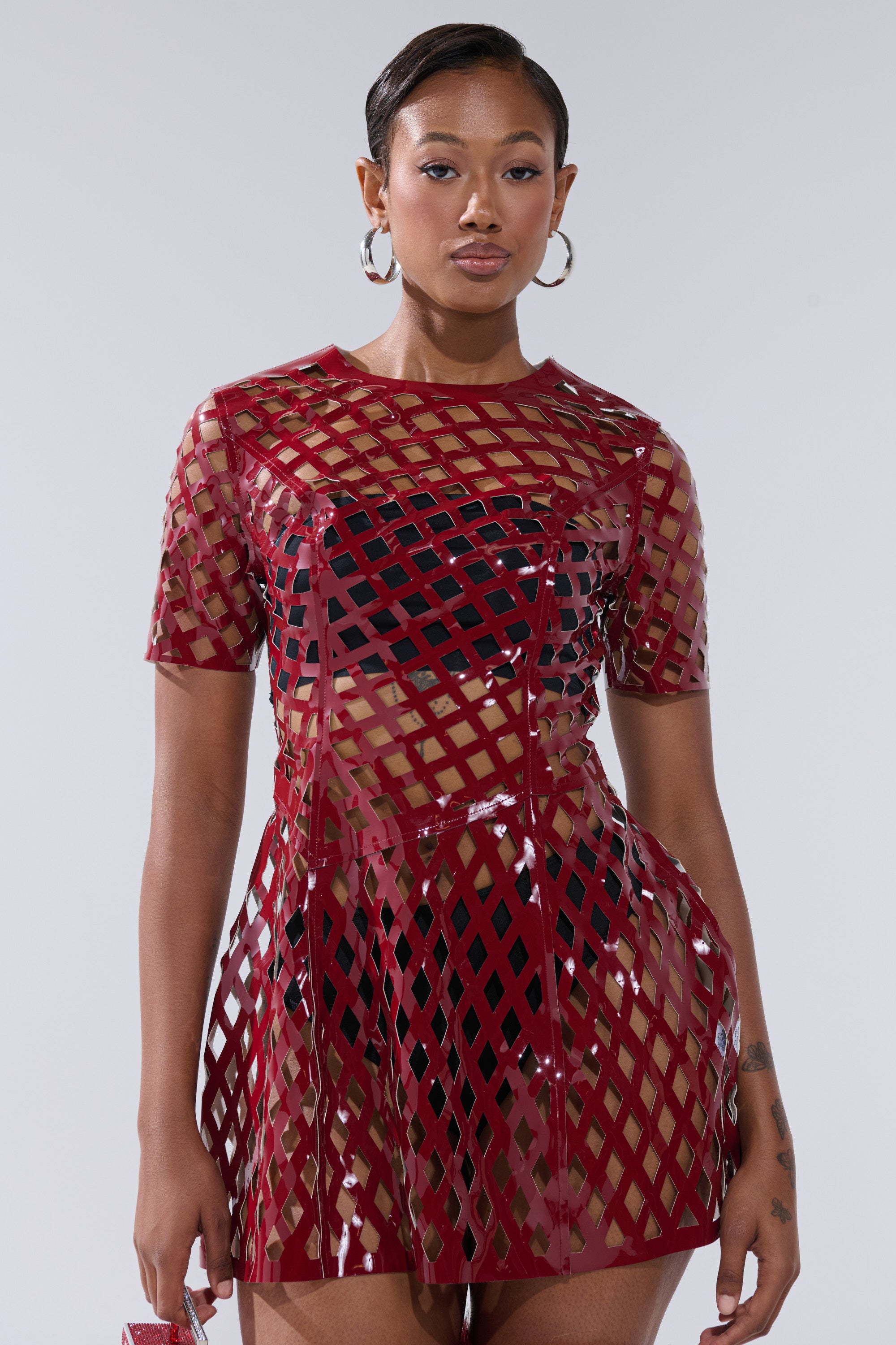 A woman with short hair and hoop earrings stands confidently against a light gray background, modeling the SHE'S DIFFERENT CUT OUT FAUX LEATHER MINI DRESS IN BURGUNDY, featuring shiny geometric cutouts over a nude underlayer.