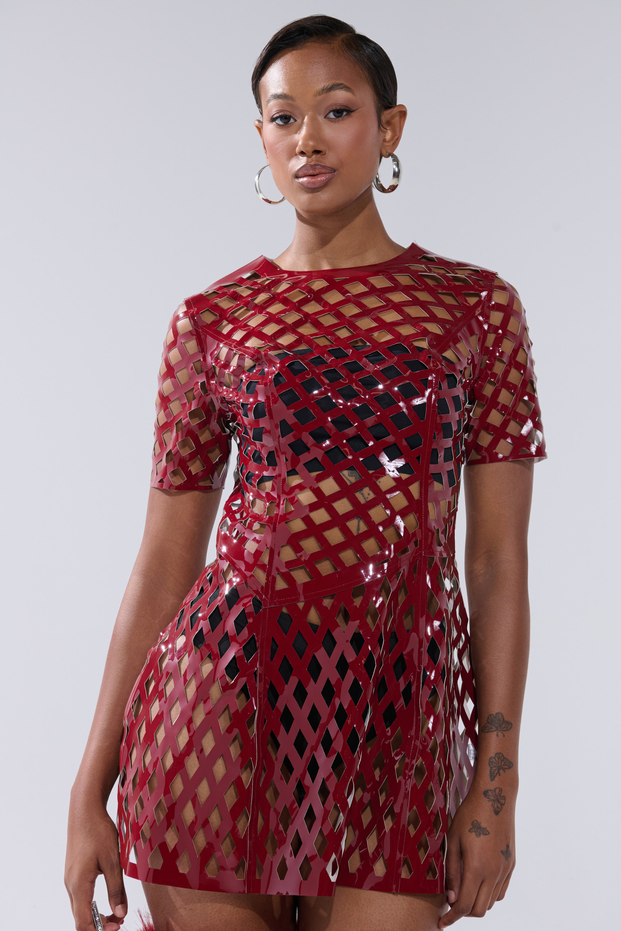 A woman poses confidently against a light gray background, wearing the SHE'S DIFFERENT CUT OUT FAUX LEATHER MINI DRESS IN BURGUNDY with large hoop earrings and visible tattoos on her left forearm.