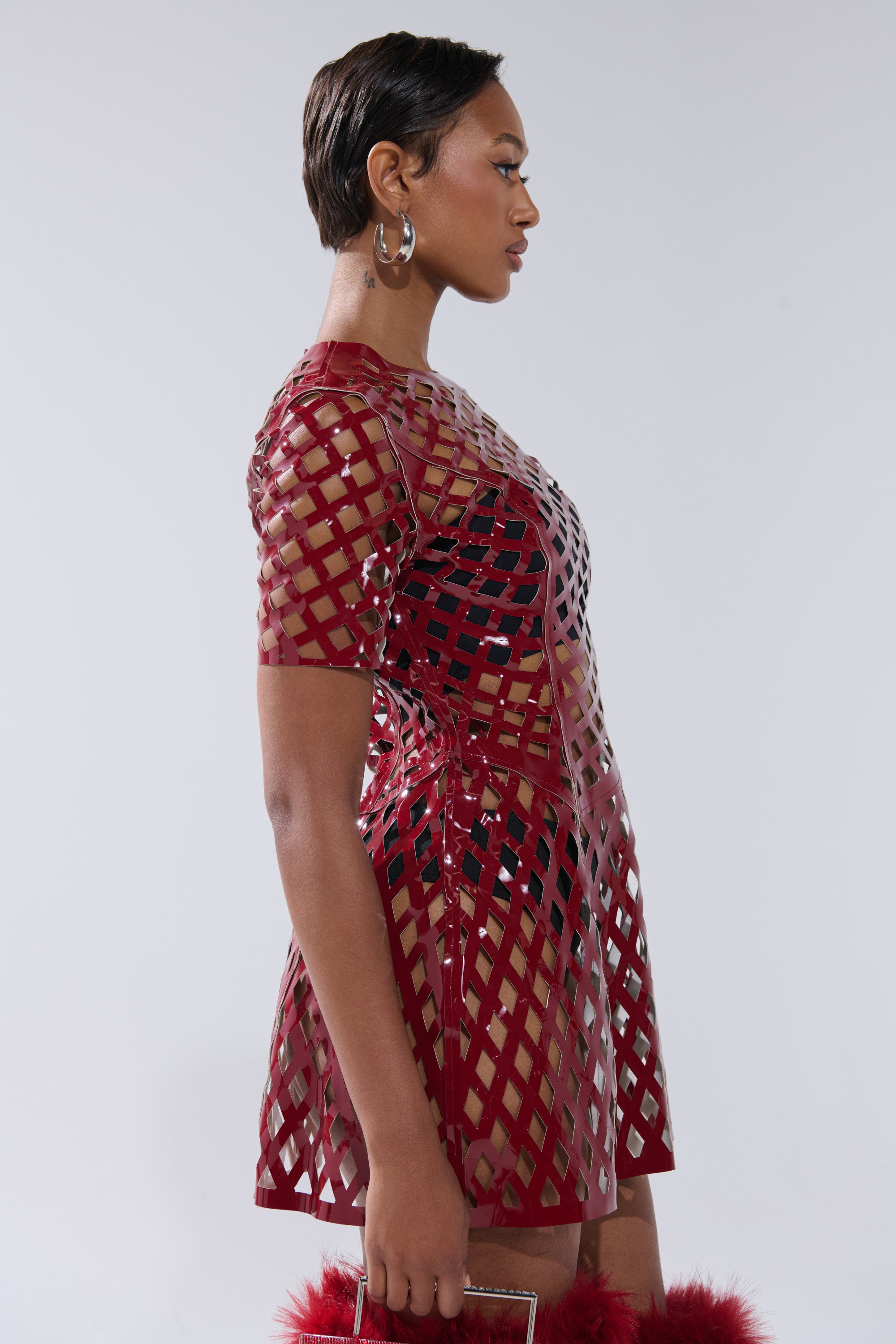 A woman in profile wears the SHE'S DIFFERENT CUT OUT FAUX LEATHER MINI DRESS IN BURGUNDY, a short-sleeved mini dress. She carries a small handbag with red fur trim, wears hoop earrings, and stands against a plain light background.