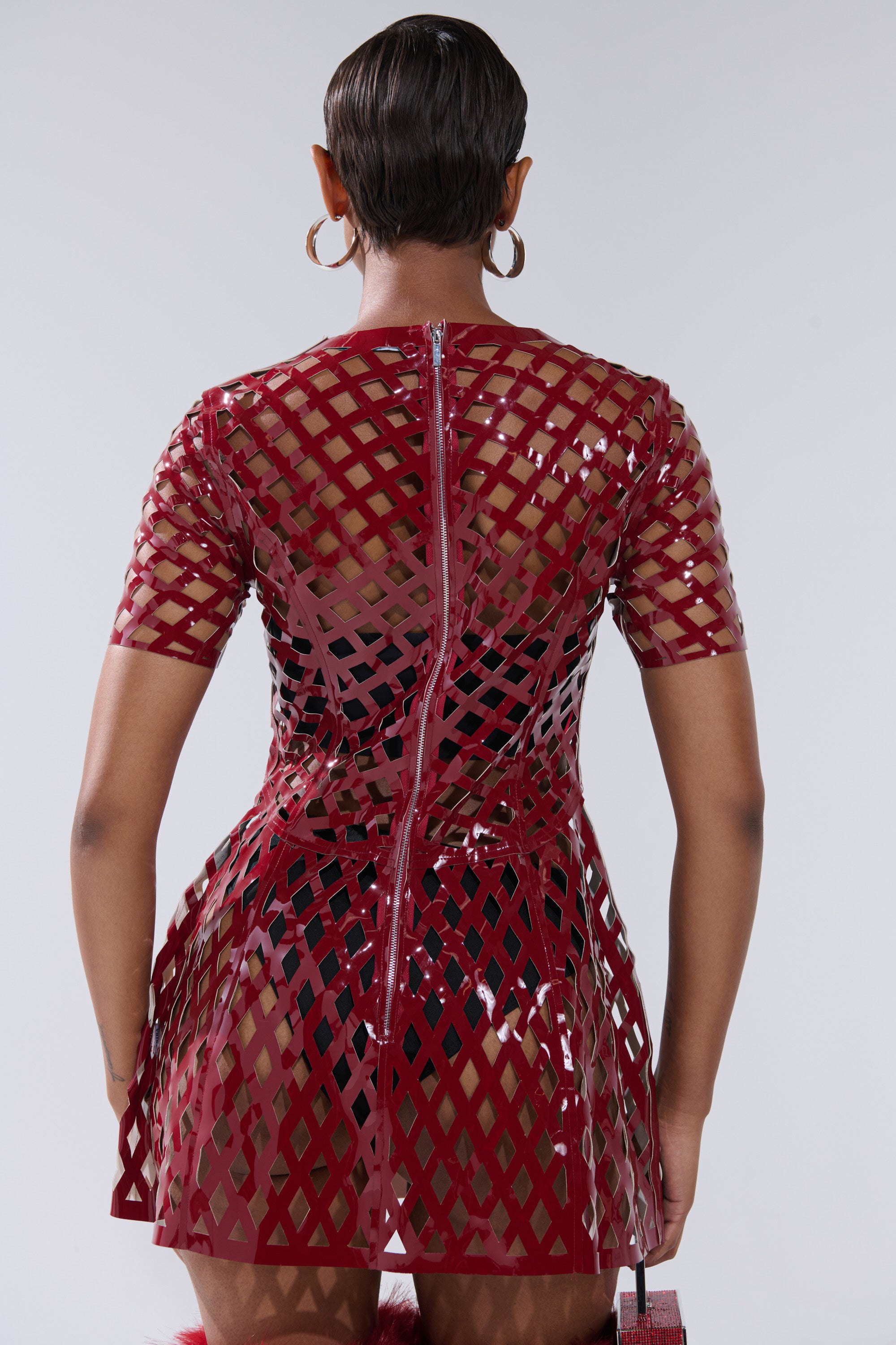 A person with short hair and hoop earrings is shown from the back wearing the SHE'S DIFFERENT Cut Out Faux Leather Mini Dress in Burgundy, featuring a zippered lattice design that reveals a black undergarment.