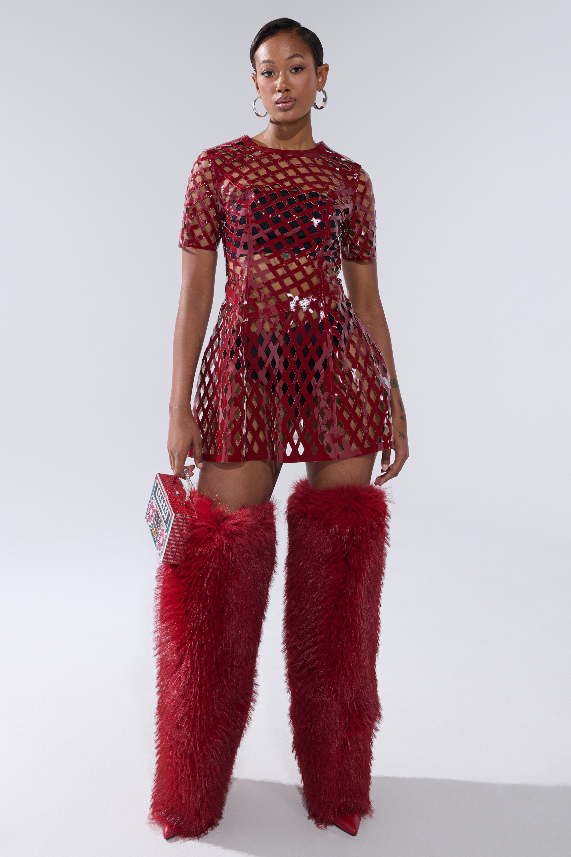 A woman stands confidently in the SHE'S DIFFERENT CUT OUT FAUX LEATHER MINI DRESS IN BURGUNDY, accessorized with a box-shaped bag and hoop earrings. She pairs the dress with tall, fluffy red boots against a plain light background.