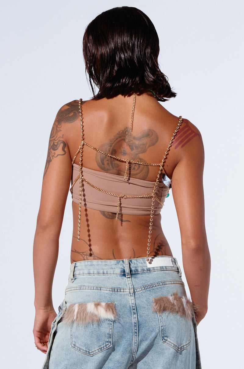 From behind, a tattooed person with short hair showcases the DON'T STOP MOVING BEADED TOP, which boasts chain details and a halter necklace. This is paired with denim pants adorned with fur accents. The overall look epitomizes festival fashion—casual and eclectic.