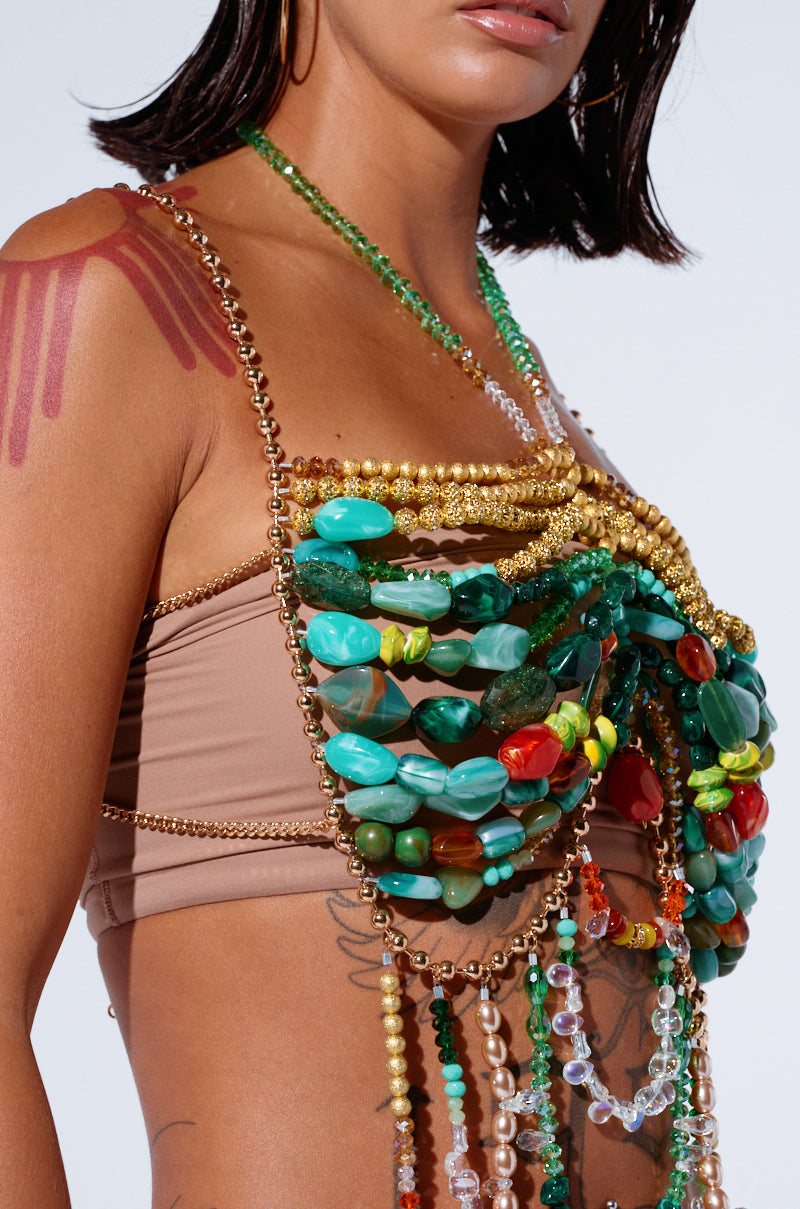 A person wearing the DON'T STOP MOVING BEADED TOP, an iridescent beaded halter necklace with green, gold, and red stones and charms draped over a tan crop top. The person's shoulder has red paint streaks, and they have dark chin-length hair. Their arm sports a tattoo, capturing the essence of festival season against a plain background.