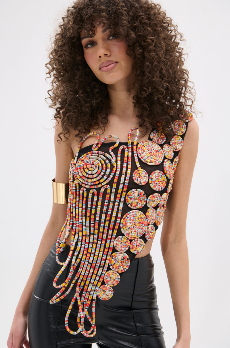 A woman with curly hair models the ROUND AND ROUND ASYMMETRIC SLEEVELESS TOP, featuring colorful beaded circular patterns, paired with a black fitted skirt and a wide metallic arm cuff, set against a plain white background.