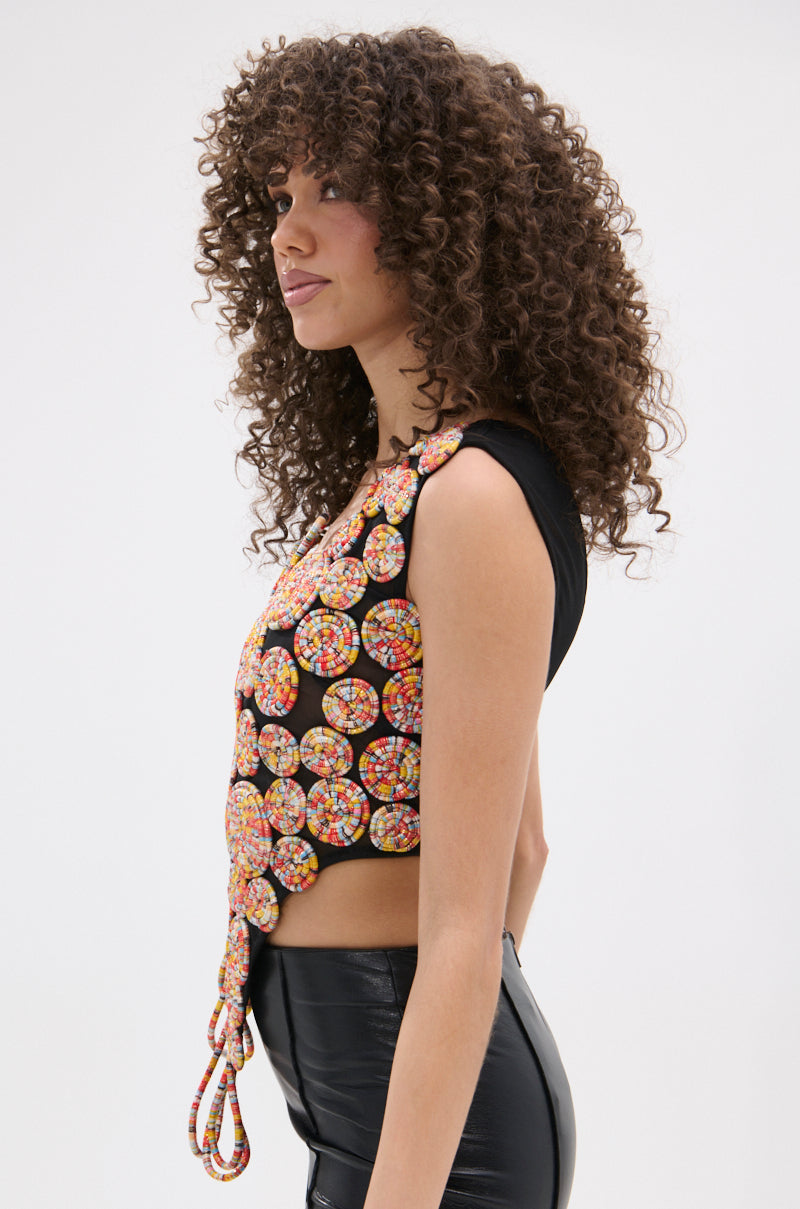A woman with curly hair stands in profile against a plain white background, wearing the ROUND AND ROUND ASYMMETRIC SLEEVELESS TOP made of colorful, circular beads over a black shirt and shiny black pants.
