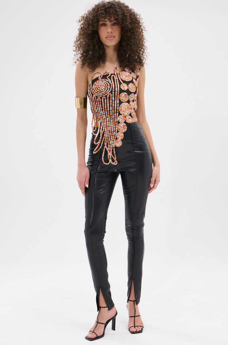 A woman with curly hair wears the ROUND AND ROUND ASYMMETRIC SLEEVELESS TOP, featuring metallic details and an abstract print, paired with shiny black high-waisted pants with front ankle slits, black heeled sandals, and a gold cuff bracelet.