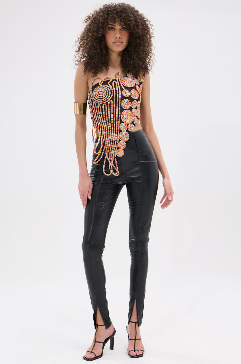 A woman with curly hair stands against a white background, wearing the ROUND AND ROUND ASYMMETRIC SLEEVELESS TOP, shiny black high-waisted pants with ankle slits, black strappy heels, and a gold arm cuff.