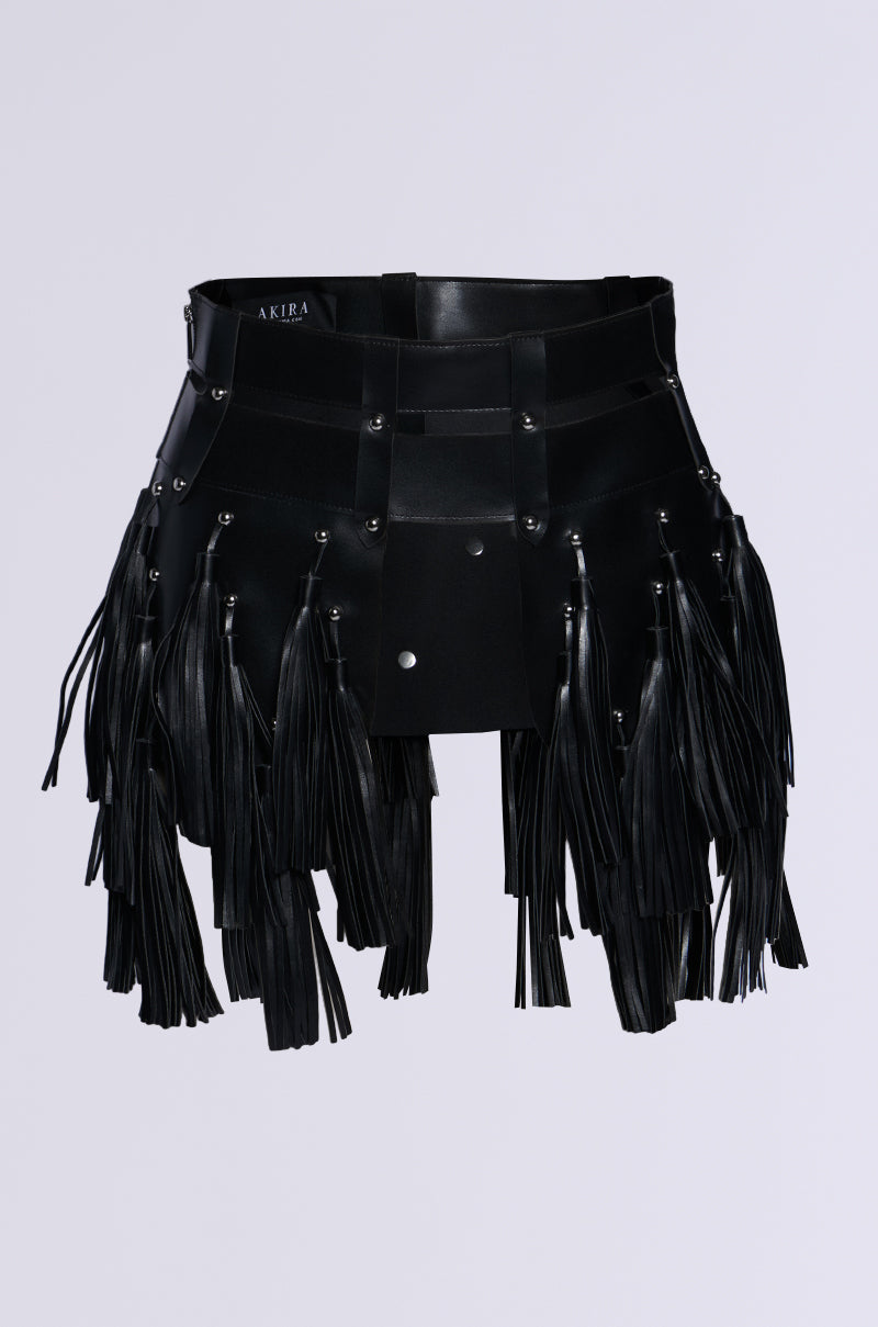 The IM WORTH IT TASSEL BELT, featuring fringe detailing and metal studs on a black faux leather mini skirt, creates a bold, grunge-chic look against a plain white background.