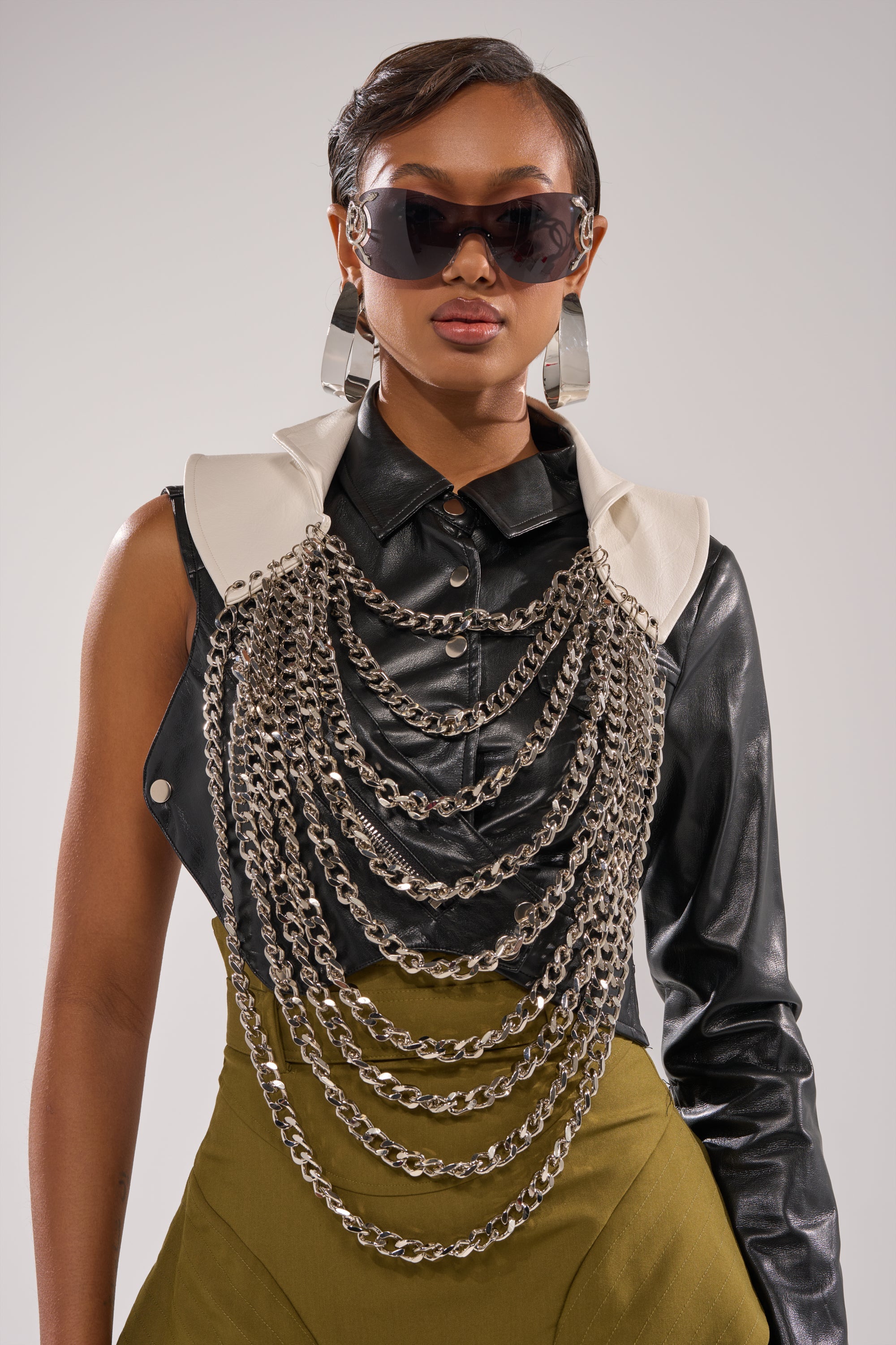 A stylish woman wears dark sunglasses, large metallic earrings, a black leather shirt with white trim, an olive-green skirt, and the HEAVY METAL NECKLACE—a bold silver chain piece that completes her statement look. 1