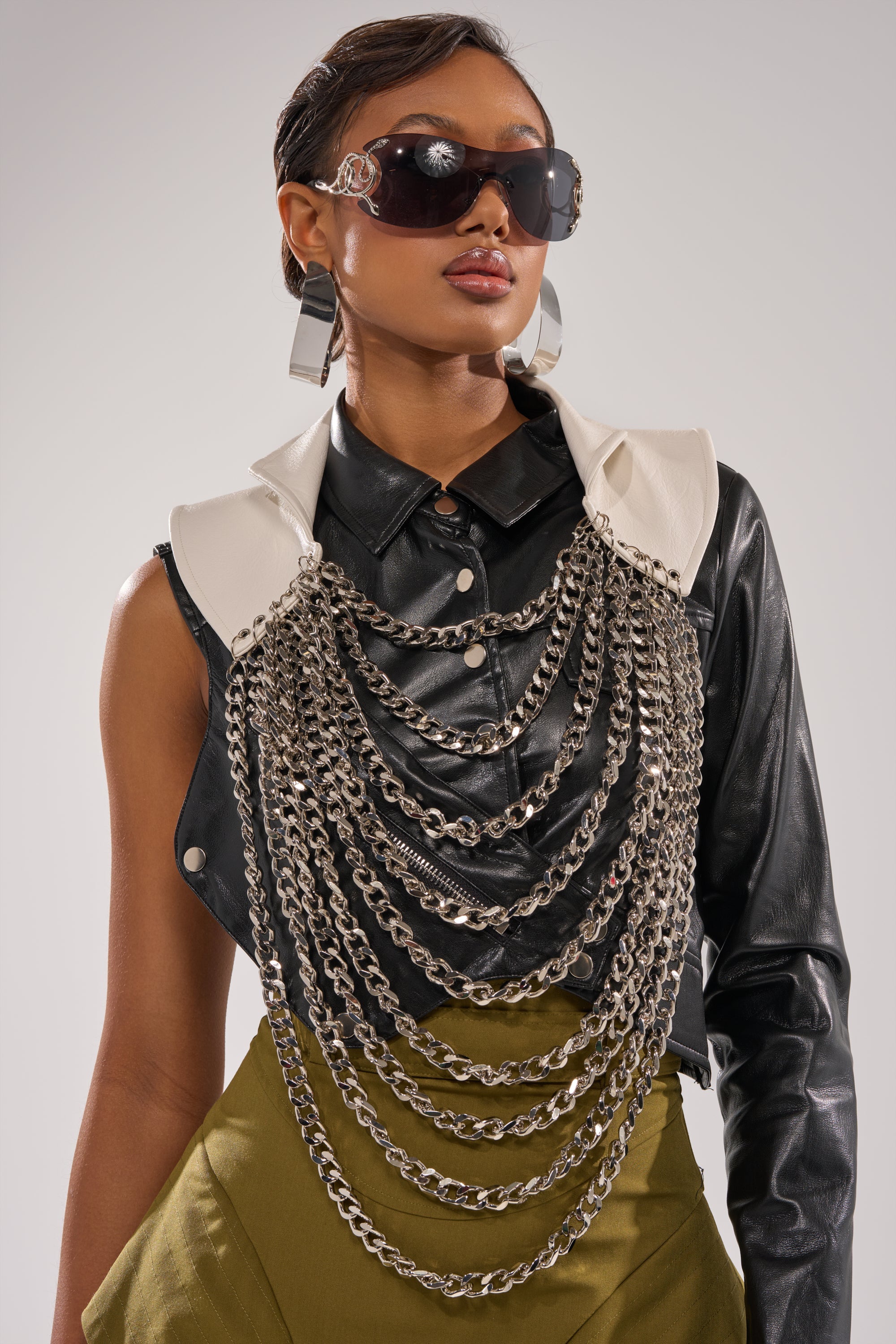 A woman with slicked-back hair wears olive green pants, a black one-sleeve leather shirt under a white shoulder accent, oversized sunglasses, large geometric earrings, and the HEAVY METAL NECKLACE as an eye-catching statement piece. 2