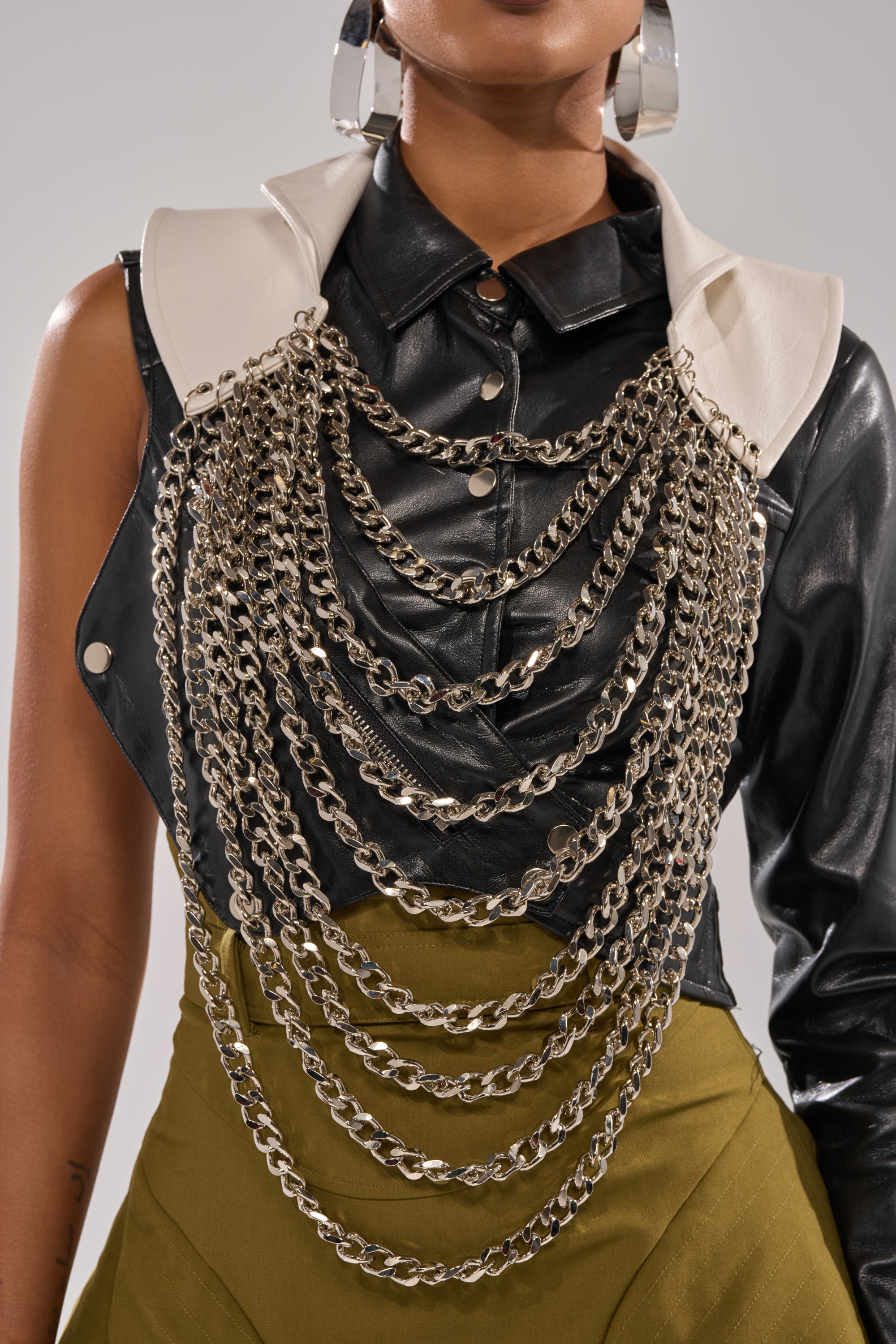 A person wears a black leather sleeveless top with a white collar, large silver hoop earrings, olive green high-waisted pants, and the HEAVY METAL NECKLACE as a bold statement piece across the chest. 3