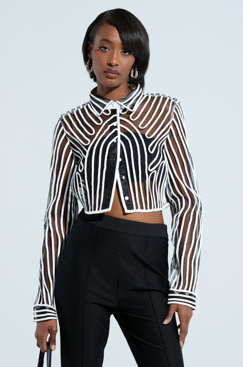 A woman wearing the EVA LONG SLEEVE BUTTON DOWN STRIPED SHIRT in black and white sheer mesh over a cropped black top and high-waisted black pants. She has short, sleek hair and is holding a small handbag against a plain, light-colored background.