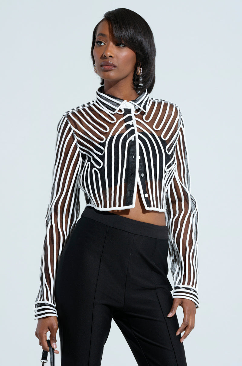 A woman stands confidently, wearing the EVA LONG SLEEVE BUTTON DOWN STRIPED SHIRT in chic black and white sheer mesh, paired with black high-waisted pants. Her straight, shoulder-length hair perfectly complements her look. She holds a small handbag and gazes off to the side against a plain background.