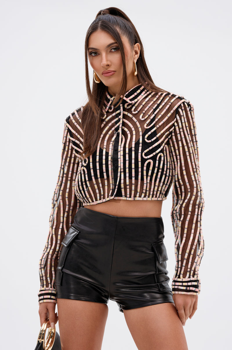 A woman wears the EVA LONG SLEEVE BUTTON DOWN STRIPED SHIRT with high-waisted black leather shorts and holds a small purse while standing against a plain white background.