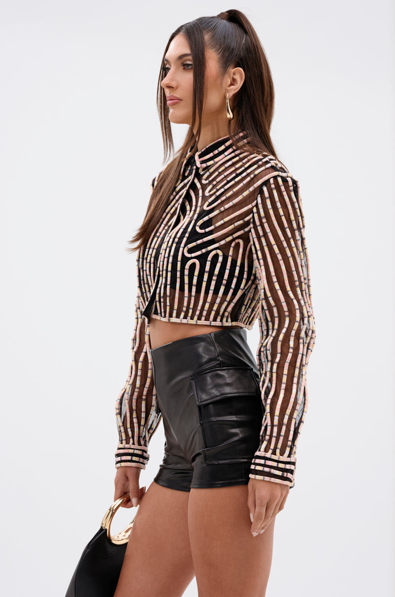 A woman stands in profile wearing the EVA LONG SLEEVE BUTTON DOWN STRIPED SHIRT with high-waisted black leather shorts. She holds a small black handbag, and her long straight hair is styled in a high ponytail.